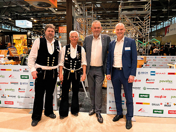 Vier Männer stehen auf einer Messe vor einer Sponsorentafel; zwei tragen traditionelle Zimmermannskleidung.