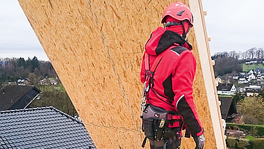 Person mit Helm, Schutzkleidung und persönlicher Schutzausrüstung gegen Absturz schiebt auf einem Dach ein hängendes Holzbauelement weg.