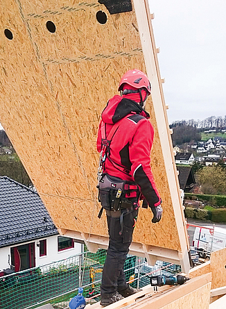 Person mit Helm, Schutzkleidung und persönlicher Schutzausrüstung gegen Absturz schiebt auf einem Dach ein hängendes Holzbauelement weg.