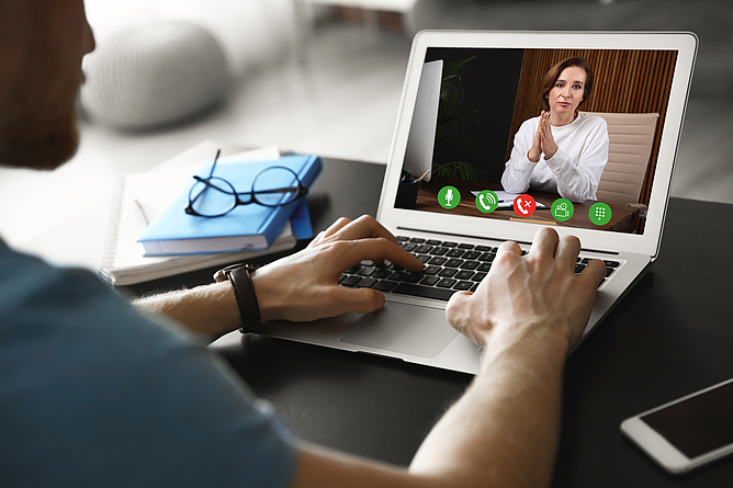 Ein Mann sitzt am Schreibtisch mit einem Laptop. Er führt eine Videokonferenz mit einer Frau. 