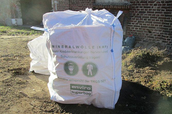 Großer weißer Sack mit der Aufschrift „Mineralwolle (KMF)“ auf einer Baustelle vor einem Backsteingebäude.