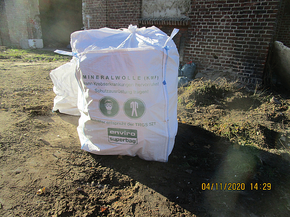 Großer weißer Sack mit der Aufschrift „Mineralwolle (KMF)“ auf einer Baustelle vor einem Backsteingebäude.