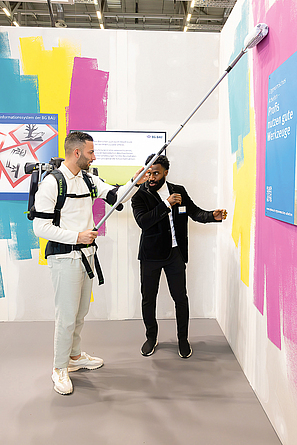 Ein Mann mit einem Exoskelett bewegt eine Teleskopstange mit Farbrolle an der Wand. Ein anderer betreut den testenden Mann.