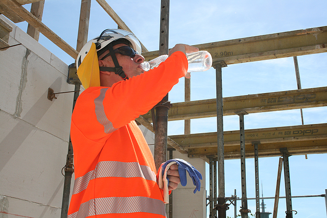Ein Bauarbeiter steht mit Schutzhelm, Nackenschutz und Arbeitskleidung auf einer Baustelle. Er trinkt bei Hitze Wasser.