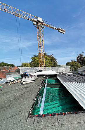 Eine ab Dachfirst geöffnete rechte Dachfläche ist mit grünen Netzen bespannt, gefolgt von Welldachplatten. Im Hintergrund befindet sich ein Kran.