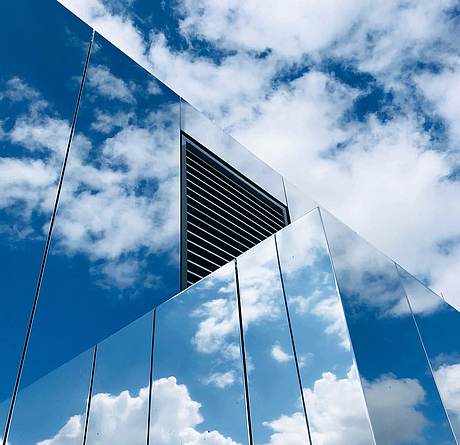 Silberner Metallbau mit Lüftungsschlitzen oben auf einem Gebäude, die Wolken spiegeln sich auf der Oberfläche der Paneele.