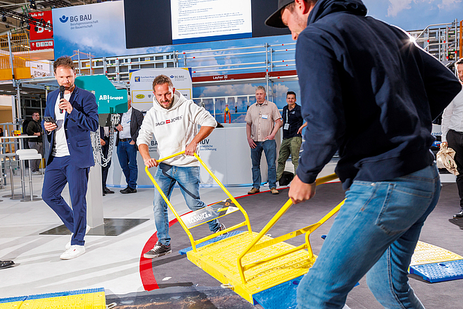 Zwei Männern stecken mit einer Verlegehilfe einzelne Module einer Grabenbrücke am Messestand zusammen. Der Mann am Rande kommentiert das Geschehen über Mikrofon.