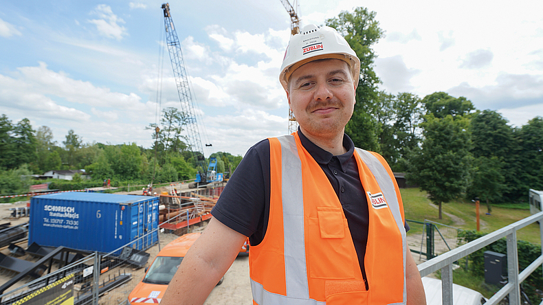 Eine Person mit Schutzhelm und orangefarbener Warnweste steht auf einer Baustelle und blickt in die Kamera.