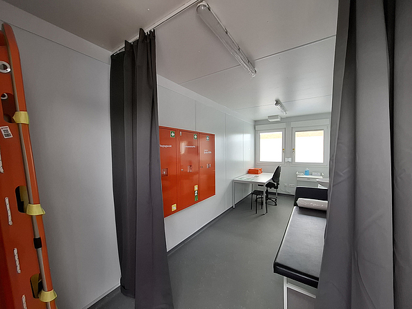 Schmaler Raum mit Liege, Tisch und Stuhl, der durch einen Vorhang abgetrennt ist. An der Wand ist eine Rettungstrage angebracht. 