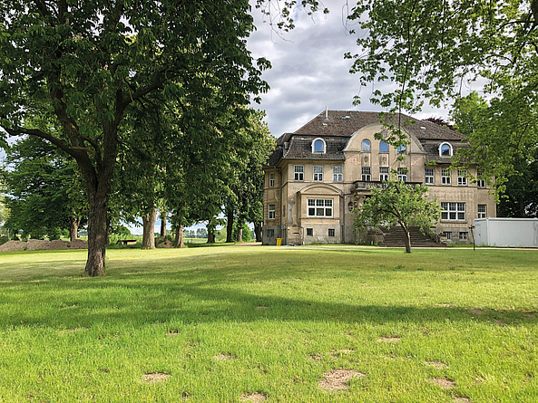 Eine große, gepflegte Rasenfläche mit mehreren Bäumen erstreckt sich vor einem historischen Gebäude.