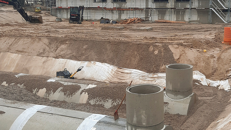 Baugrube mit eingebauten Sickertunneln und aufgesetzten Schächten aus Stahlbeton vor einem eingerüsteten Gebäude.