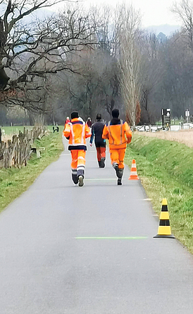 Blick von hinten auf mehrere Personen, die sich auf einer kleiner Landstraße bewegen. Die Personen haben unterschiedlich leuchtende orange-rote Warnkleidung. an. 