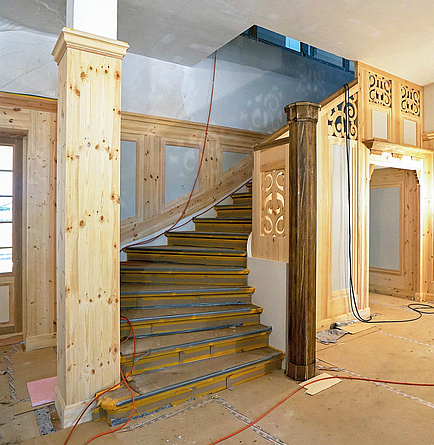 Ein Treppenaufgang aus hellem Holz in einem denkmalgeschützten Gebäude wird renoviert. Geländer und Wandverkleidungen sind teilweise erneuert.